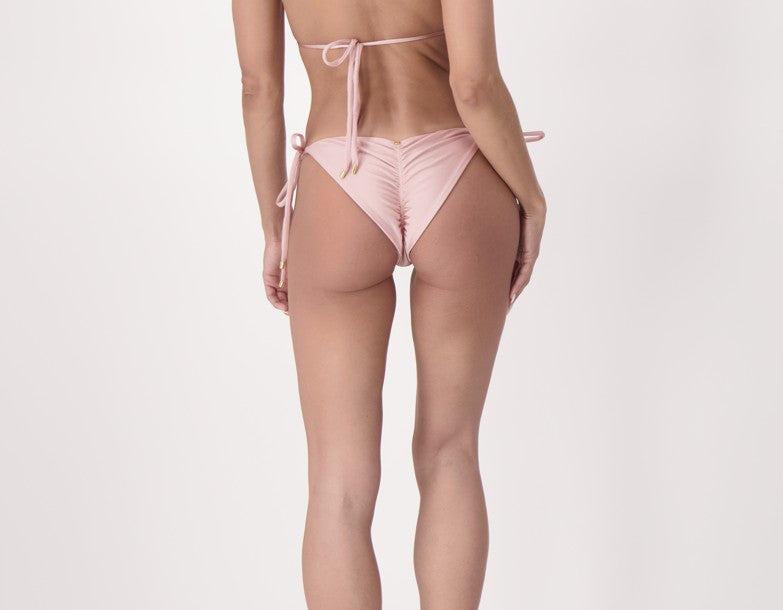 Woman wearing Cariño luxury pink satin side-tie bikini bottoms with ruched back for sculpted fit and Colombian emerald accent